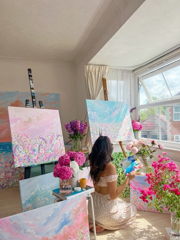 Artist painting vibrant floral canvases in a sunlit room surrounded by flowers and colorful artwork.