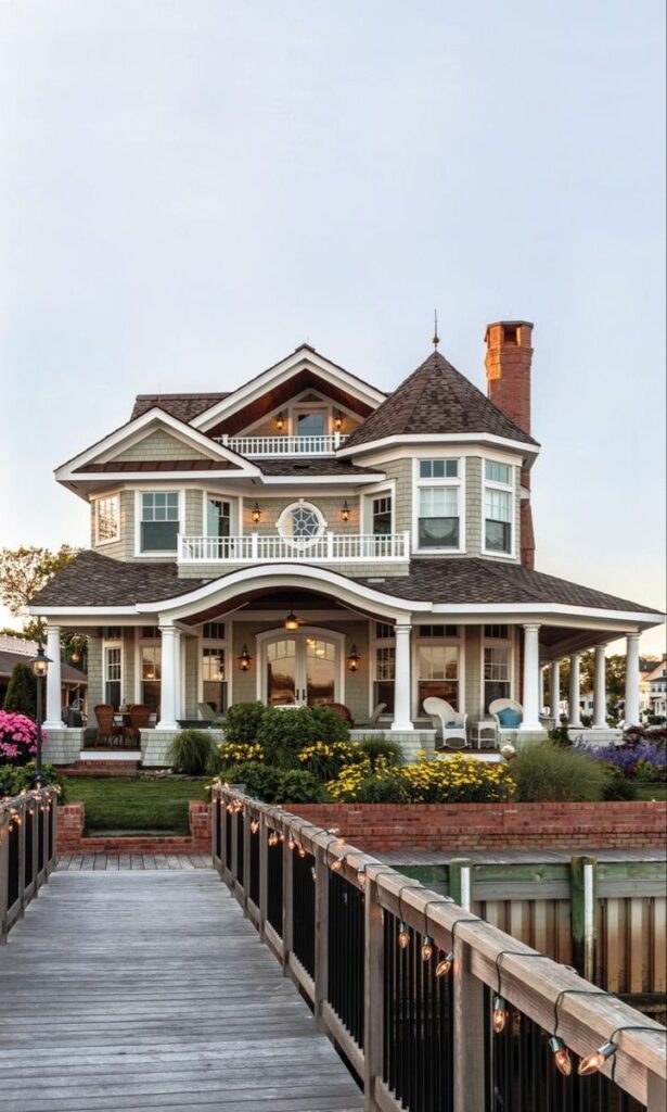 Victorian-style house with wraparound porch, tall chimney, and colorful garden, viewed from a wooden pathway adorned with lights.