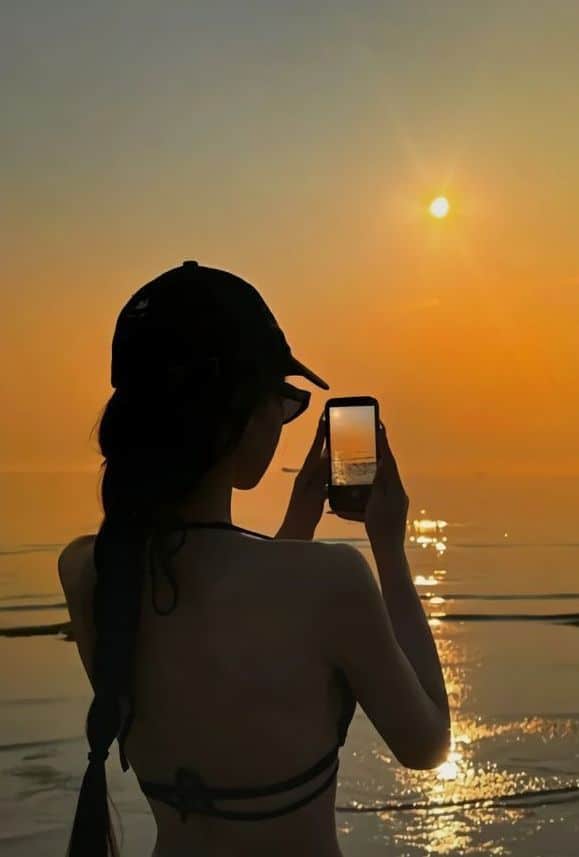 Person capturing stunning sunset on phone at the beach, with the sun casting a golden glow over the serene ocean water.