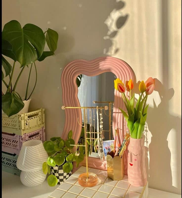 Cozy vanity setup with tulips, pink mirror, gold jewelry stand, and plants on a tiled table. Bright and stylish home decor.