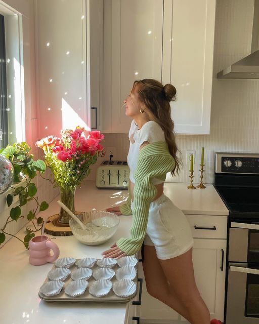 Young woman baking in a sunlit kitchen, preparing cupcakes, surrounded by pink flowers and green plants. Cozy, bright interior.