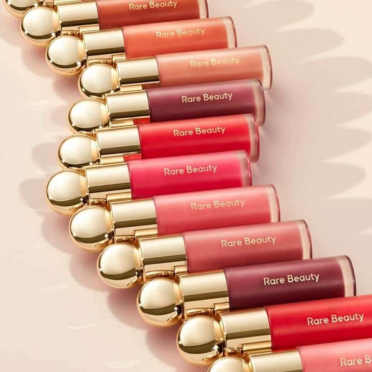 Rare Beauty lipsticks in a variety of shades with elegant gold caps, neatly arranged on a soft pink background.