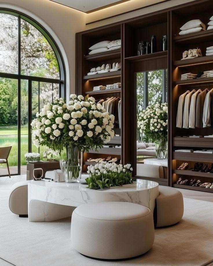 Luxurious walk-in closet with elegant decor, featuring white flowers, plush seating, and a large window overlooking greenery.