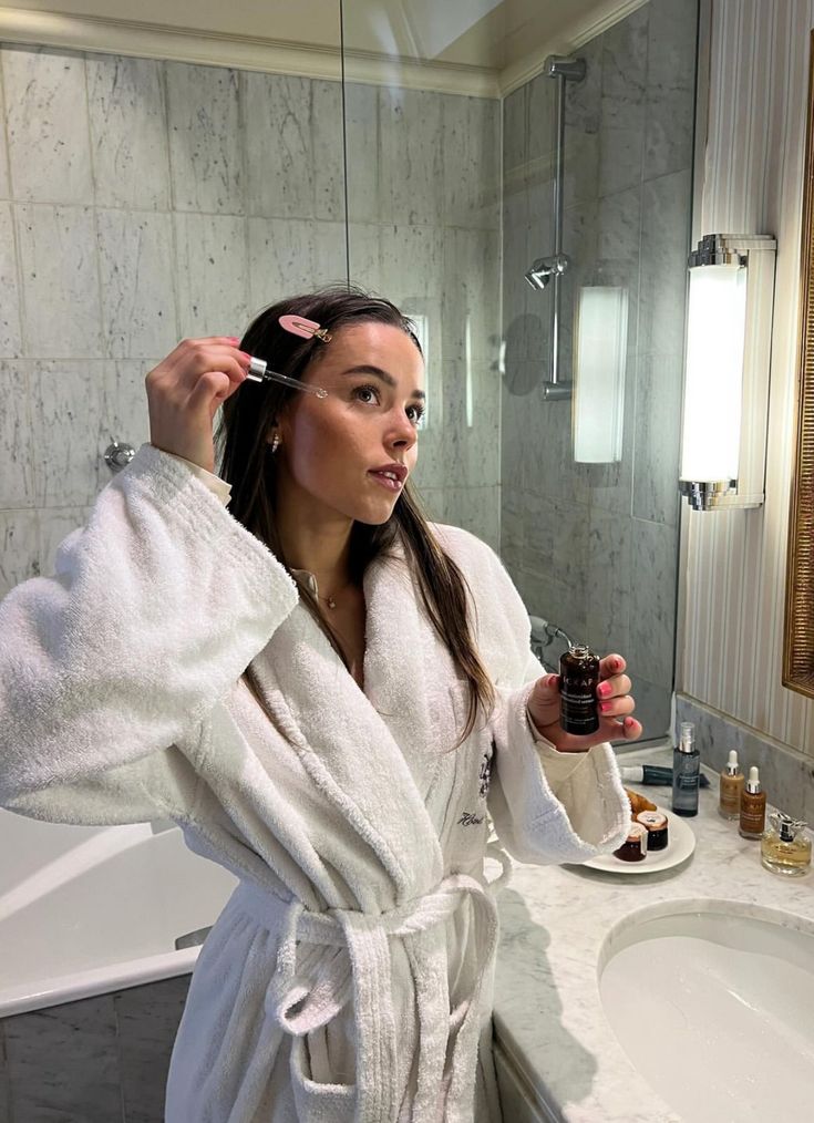Woman in a white robe applying skincare serum in a bathroom with various cosmetic bottles on the counter.