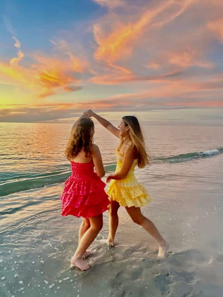 Two women dancing on the beach at sunset, wearing vibrant dresses, with colorful skies and ocean waves in the background.