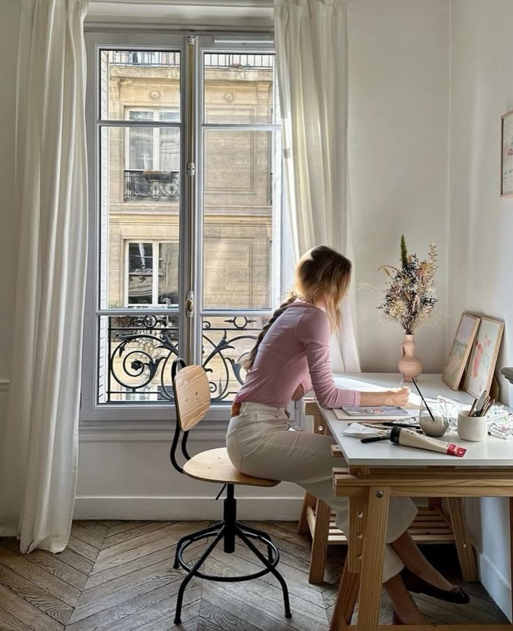 Woman working at a stylish desk by a window, with natural light and a view of classic architecture outside.