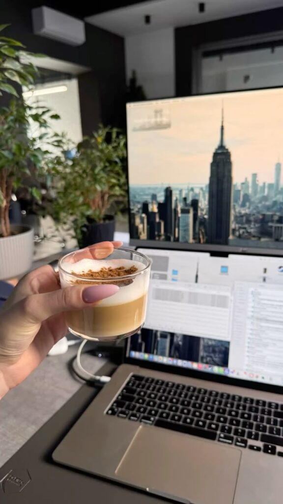 Hand holding coffee cup in front of laptop with New York skyline on screen, plants in background. Cozy office vibe.