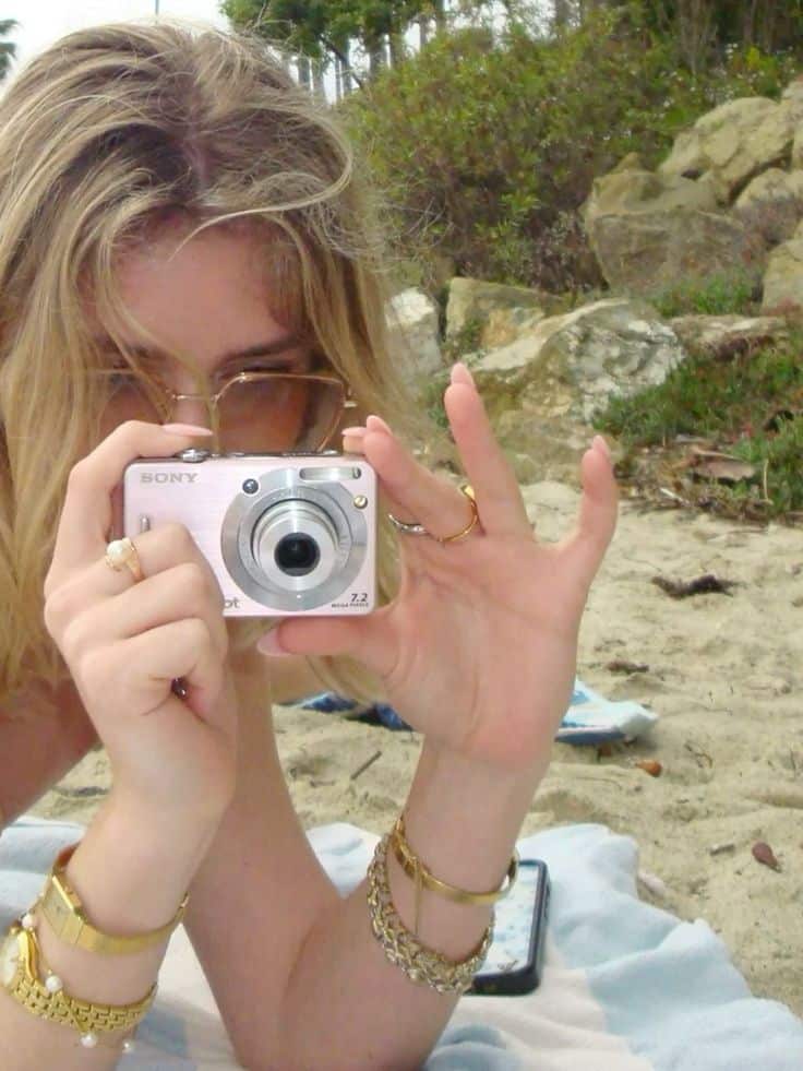 Person holding a digital camera on a beach, capturing a moment with a backdrop of sand and foliage.