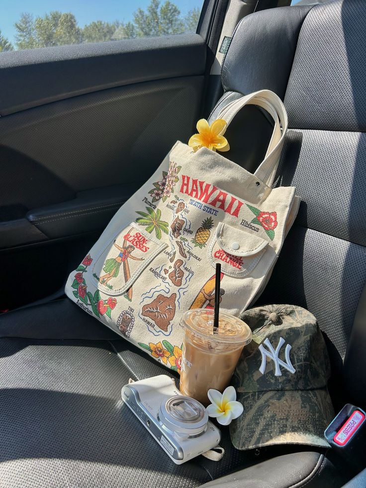 Tote bag with Hawaiian print, iced coffee, camera, and cap on car seat, adorned with yellow flowers. Perfect travel vibe.