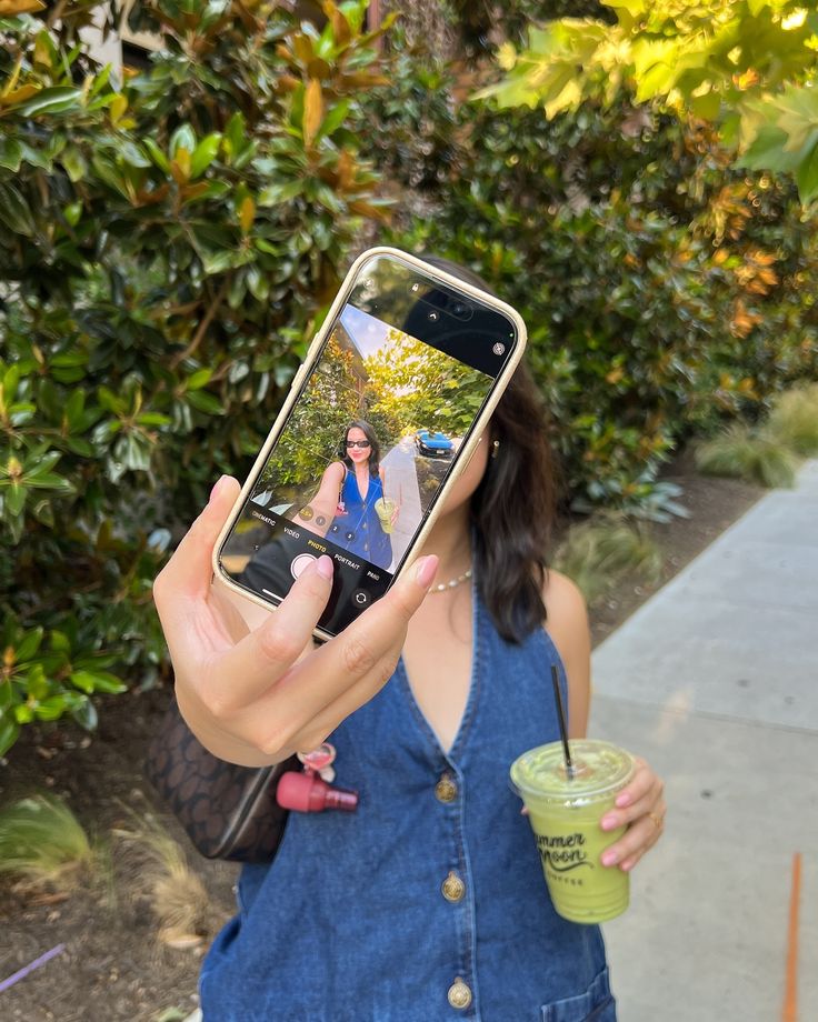 Person taking a selfie with a smartphone, holding a green drink, outdoors with leafy background. Perfect for summer vibes.
