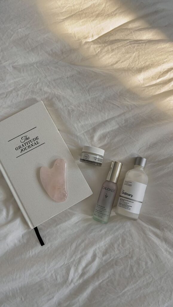 Gratitude journal, skincare products, and gua sha on a cozy white bed. Relaxation and self-care essentials flatlay.