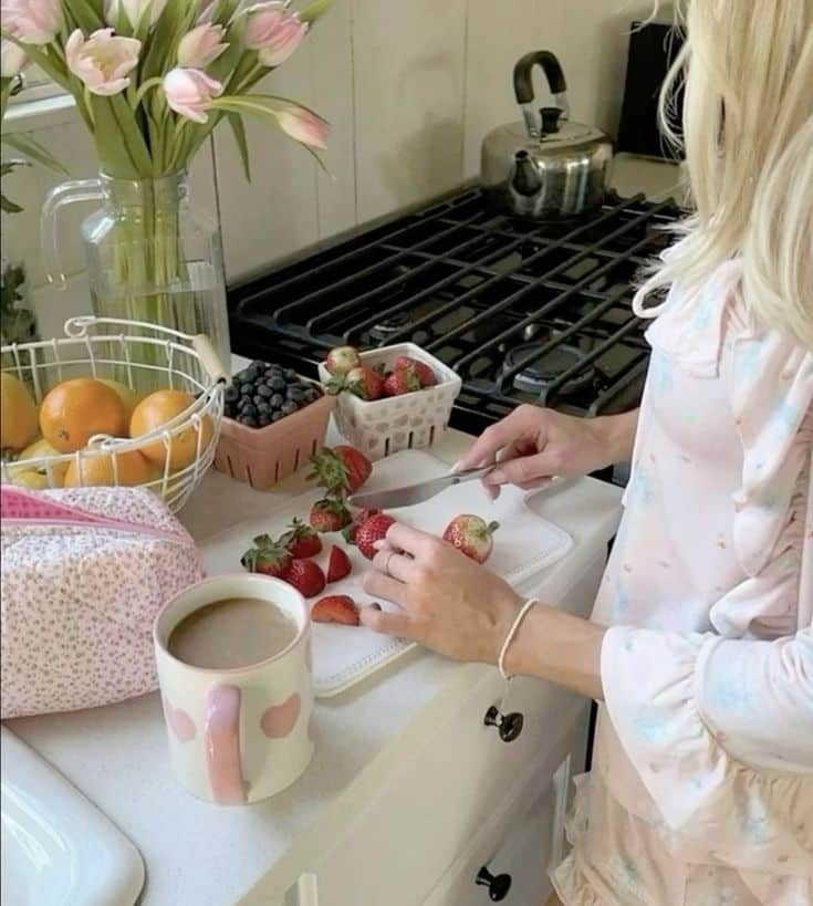 Woman in cozy pajamas slicing fresh strawberries in a kitchen, with a cup of coffee and a fruit basket nearby.