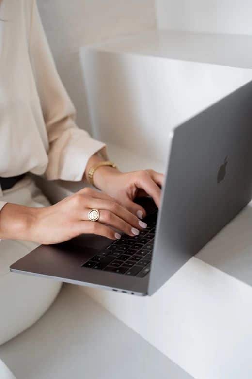 Person typing on a laptop while sitting on white stairs, wearing a gold ring and bracelet.