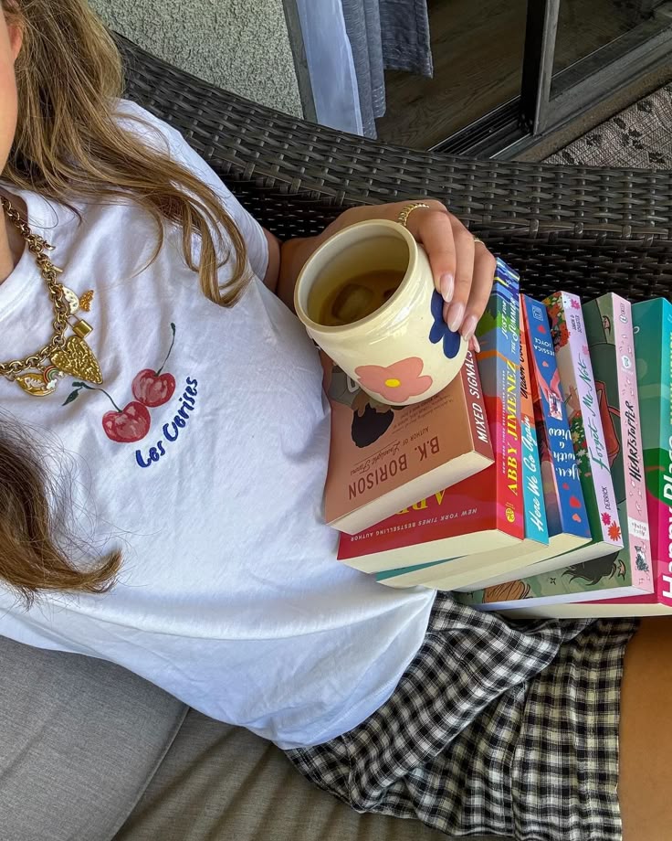 Person relaxing with tea and a stack of colorful books, wearing a cherry print t-shirt and checkered shorts. Cozy reading vibe.