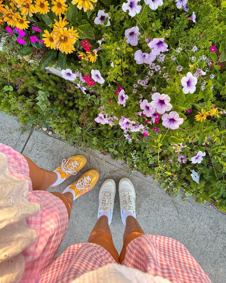 Two people in summer outfits, wearing sneakers, standing on a sidewalk beside colorful flowers. Bright, sunny day.