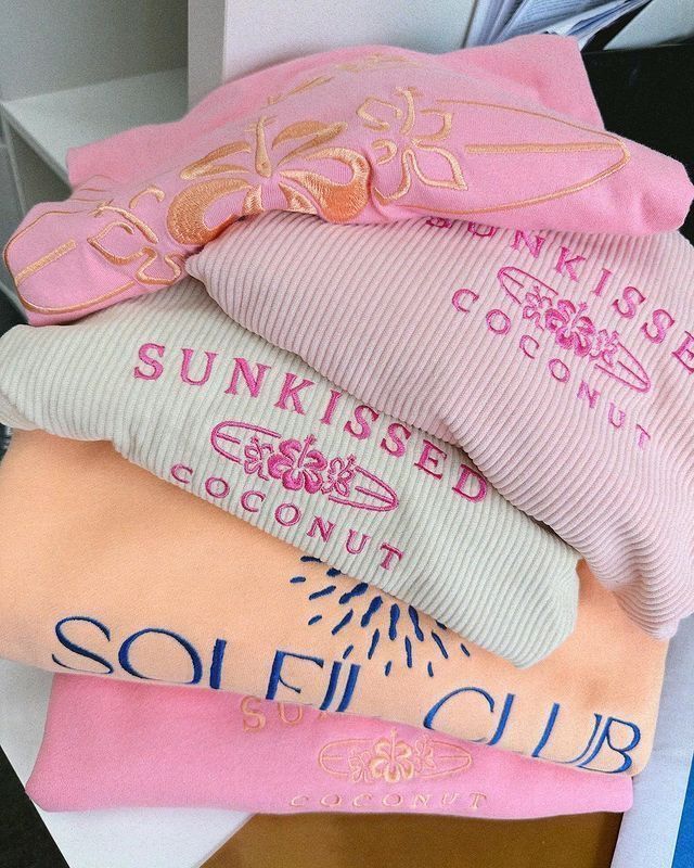 Stack of colorful sweatshirts with beach-themed embroidery, featuring text Sunkissed Coconut and Soleil Club.