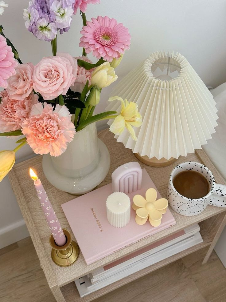 Cozy bedside setup with fresh flowers, candles, a pleated lamp, and a speckled mug, creating a warm, inviting atmosphere.
