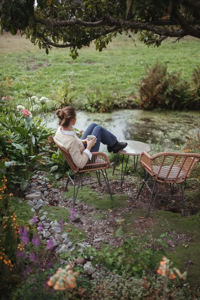 Woman relaxing with a cup of coffee by a pond, seated on a rattan chair in a lush garden. Peaceful outdoor scene.