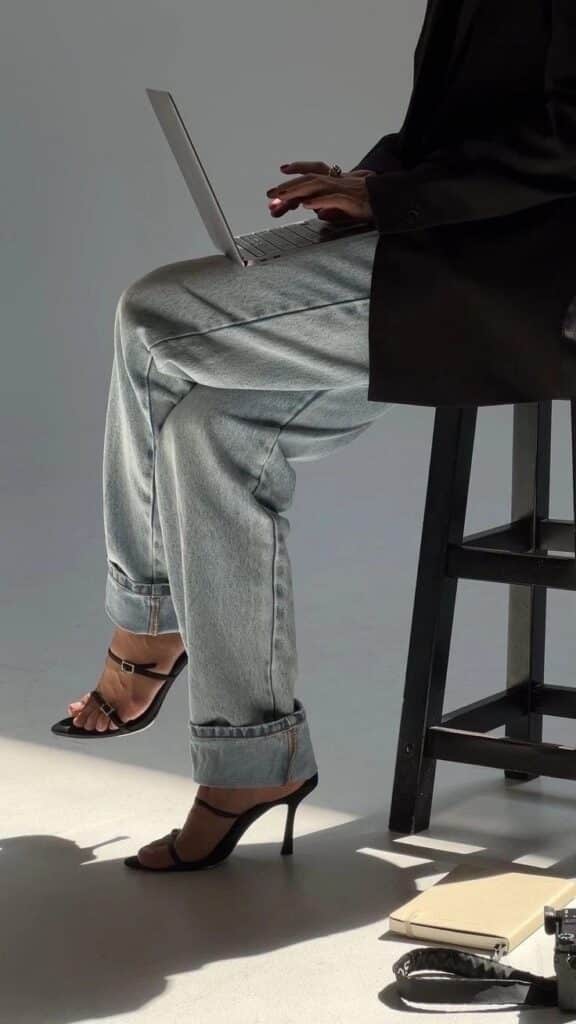 Person in jeans and heels using a laptop on a stool, with books and camera on the floor. Bright, minimalist setting.