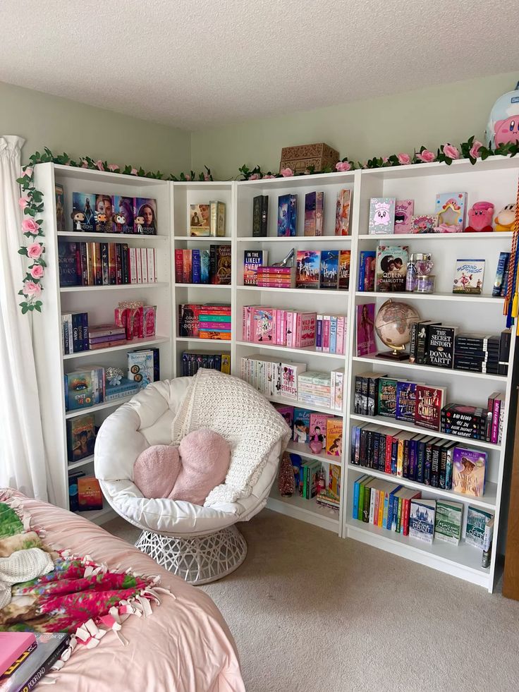 Cozy reading nook with white bookshelves filled with colorful books, a plush chair with pillows, and floral decorations.