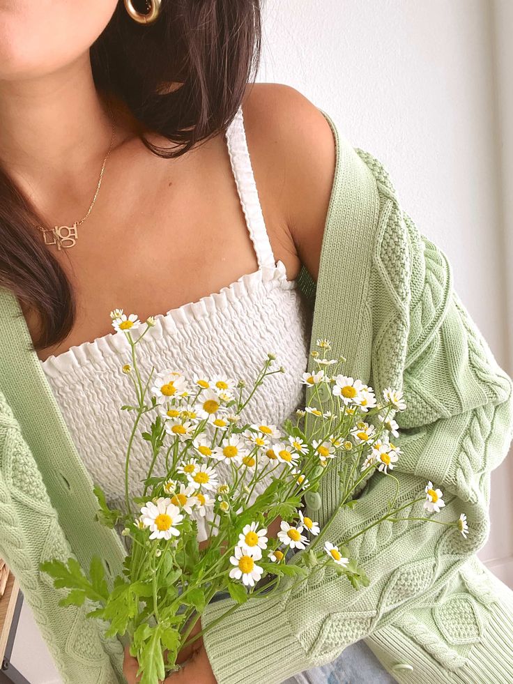 Woman in a green cardigan holding daisies, wearing a white ruched top and gold earrings. Minimalistic and fresh spring style.
