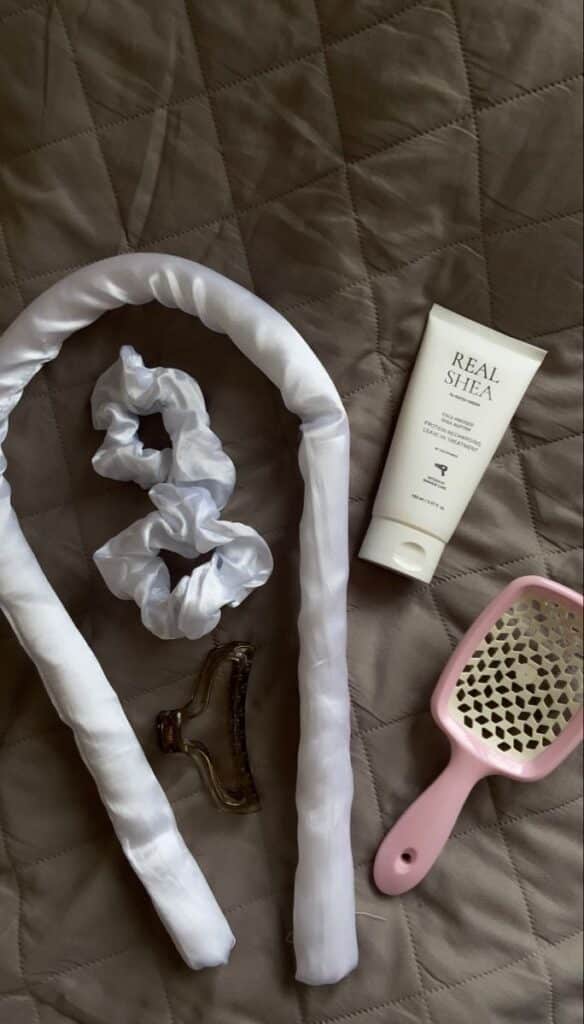 Haircare essentials on a quilted surface: silk wrap, scrunchie, claw clip, conditioner, and vented brush. Perfect for a natural hair routine.