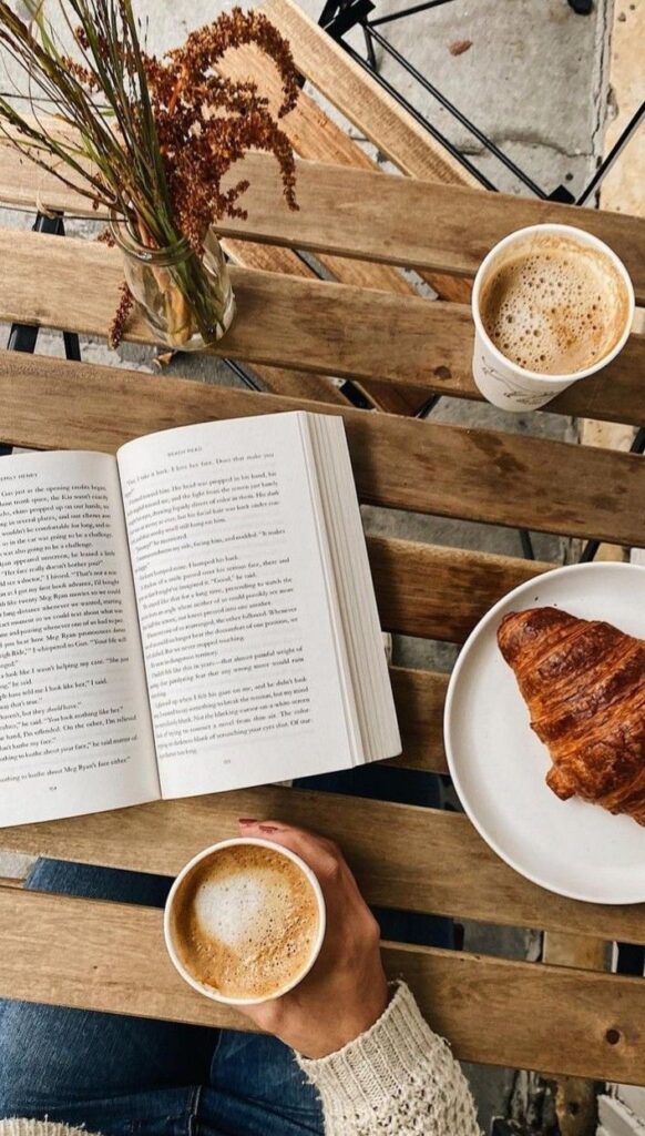 Cozy cafe scene with open book, latte, croissant, and vase of flowers on a rustic wooden table, perfect for a relaxing morning.