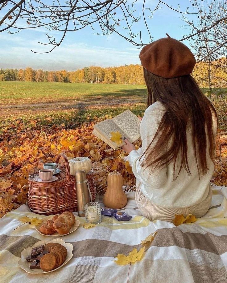 Woman enjoys autumn picnic with book and snacks on a blanket surrounded by colorful leaves and scenic landscape.