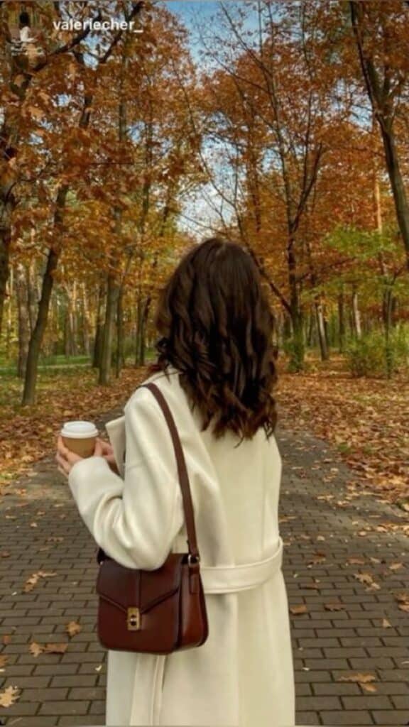 Woman in a white coat holding coffee, walking through an autumn park with colorful leaves on the ground.