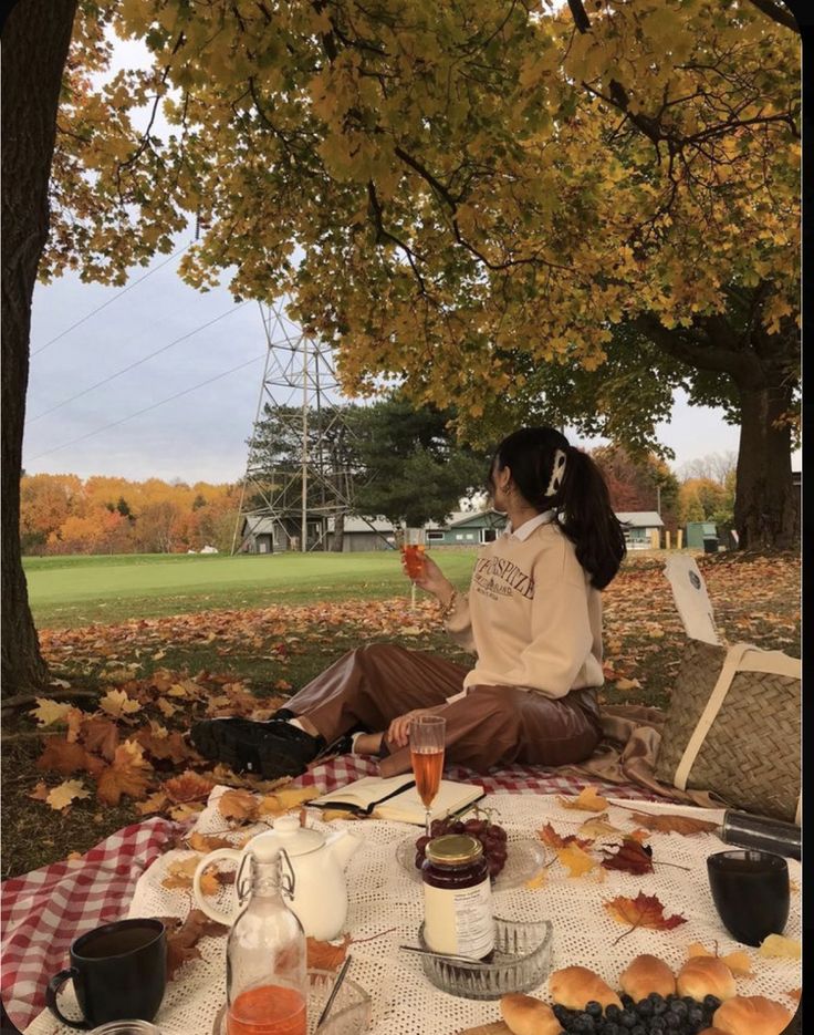 Cozy fall picnic under autumn trees with food, drinks, and a tranquil view. Woman enjoying an outdoor moment.
