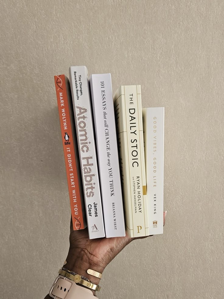 A hand holds a stack of personal development books against a beige background, showcasing titles on habits, essays, and stoicism.