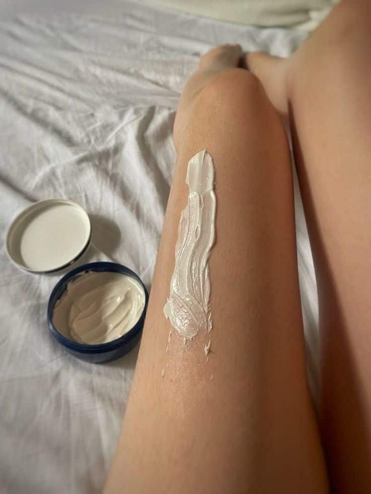 702f3faf-9e66-4056-bc7a-0f32dbaa126e - Handy Home Applying moisturizing cream on leg for smooth, hydrated skin. Skincare routine with open cream jar on bed.