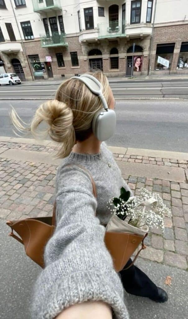 Woman with headphones, gray sweater, holding flowers in a bag, taking a selfie on a city street with historic buildings.