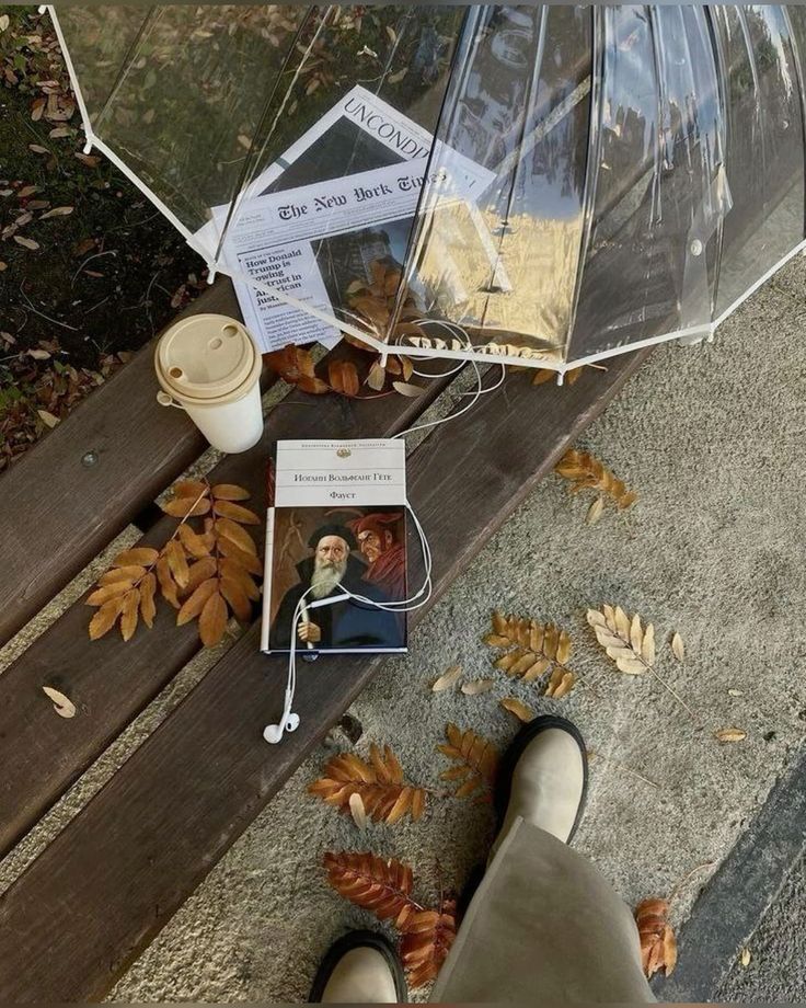 Bench with a transparent umbrella, book, newspaper, and coffee cup on a fall day, surrounded by fallen leaves and earbuds.
