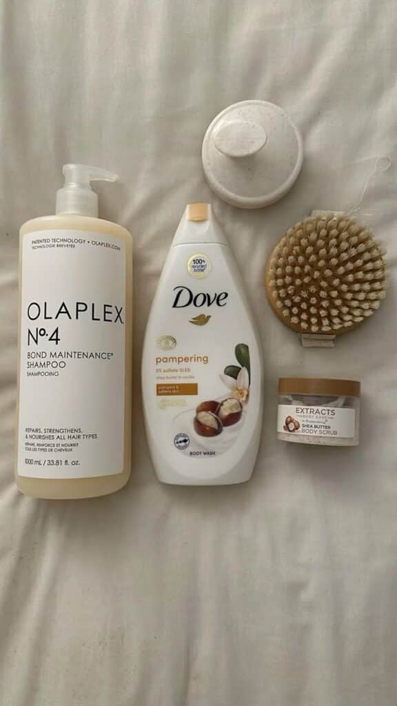 Bath essentials including Olaplex shampoo, Dove body wash, scrub brush, and shea butter body scrub on a white fabric.