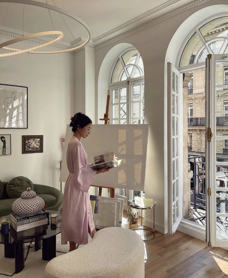 Woman in a lavender robe reading by a bright window in an elegant room with modern decor and an easel.
