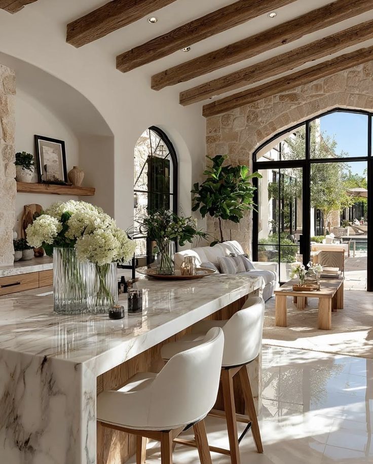 Screenshot - Handy Home Luxurious open-plan living room with marble island, wooden beams, and large windows overlooking a sunny patio garden.
