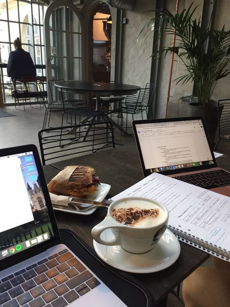 Cozy café workspace with laptops, a notebook, cappuccino, and a sandwich, ideal for remote work or study in a relaxing environment.