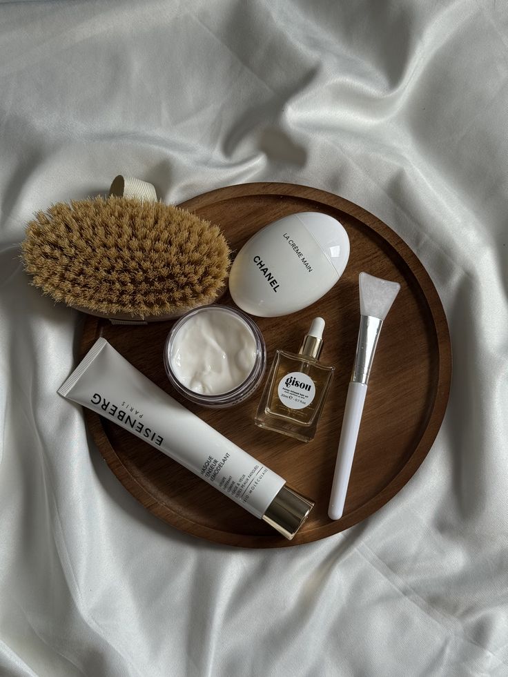 Skincare essentials on a wooden tray with soft fabric background: brush, creams, serum, and applicator brush. Luxurious self-care.