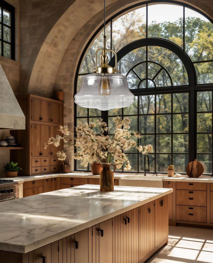 Spacious kitchen with large arched windows, wooden cabinets, marble island, and pendant light, featuring vase of flowers.