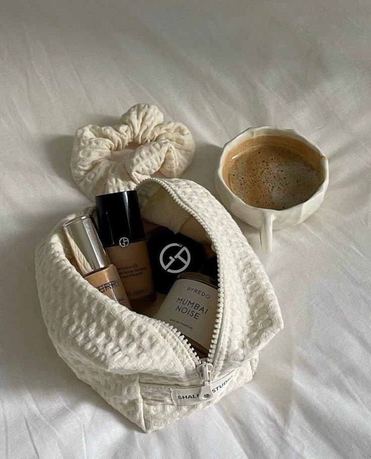 Makeup essentials and coffee on bed: foundation, perfume, lipstick in a textured pouch, cozy morning vibe.
