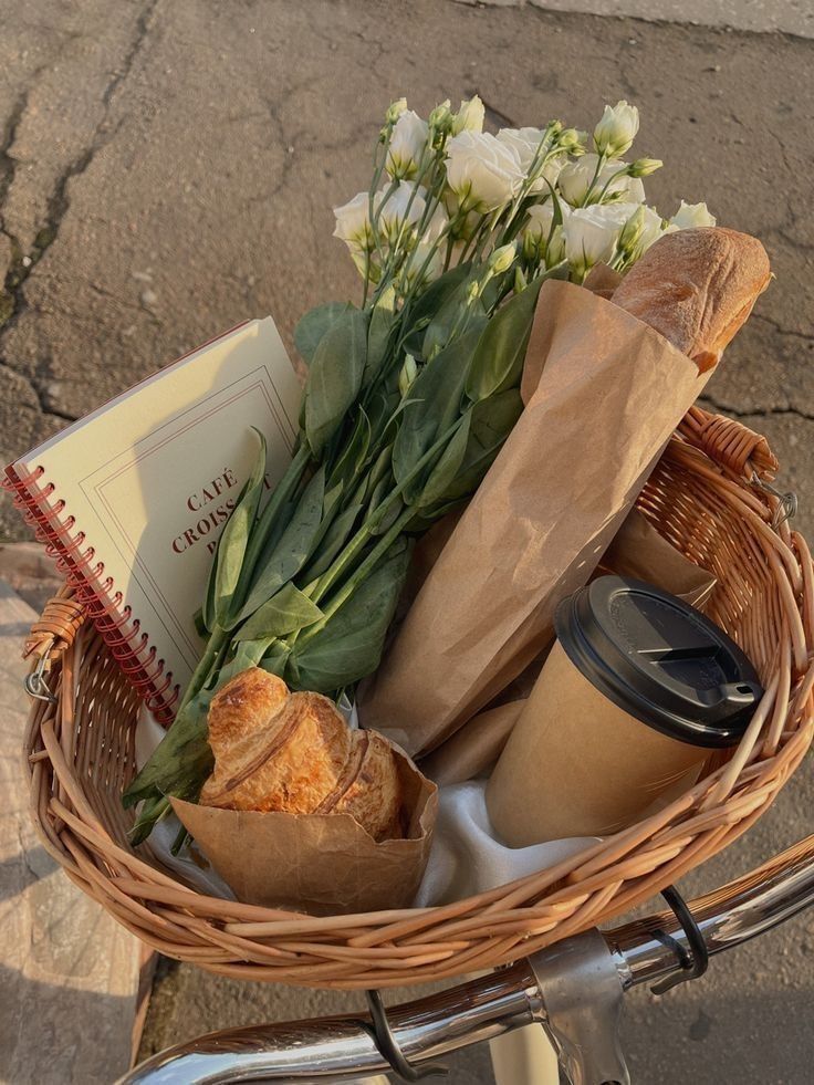 Bicycle basket with fresh flowers, baguette, croissant, coffee cup, and café menu on a sunny day. Perfect for a French picnic vibe.