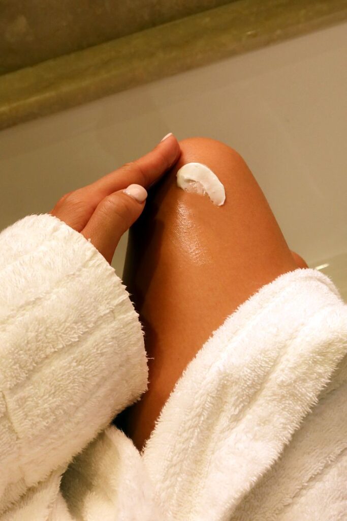 Woman applying moisturizer to leg while wearing a white robe, promoting skincare and self-care routine.