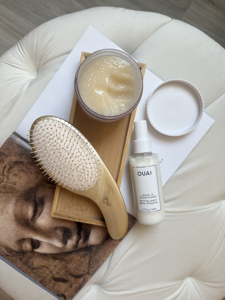 OUAI leave-in conditioner and hair brush on a tray with a jar of cream, set on a decorative book and a plush chair.