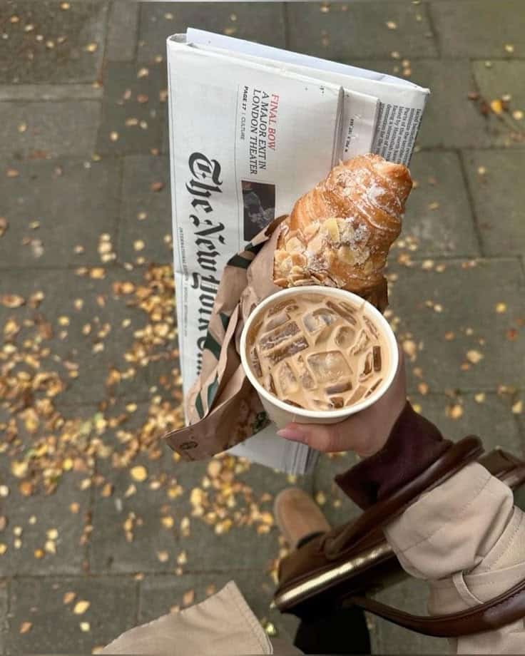 Person holding iced coffee, croissant, and newspaper on a leaf-strewn sidewalk, capturing a cozy autumn morning vibe.