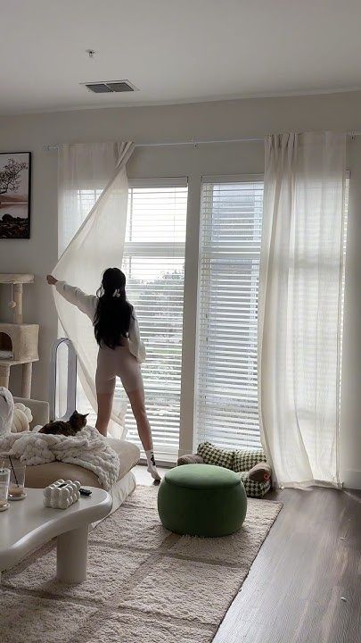 Woman opening curtains in a cozy living room, sunlight streaming in, with a cat on the sofa and a dog on the floor.