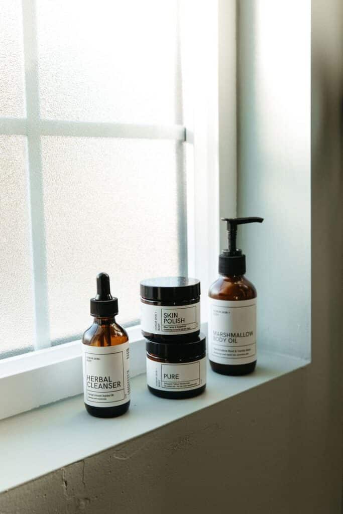 Skincare products on a window ledge, featuring herbal cleanser, skin polish, and marshmallow body oil in amber glass bottles.