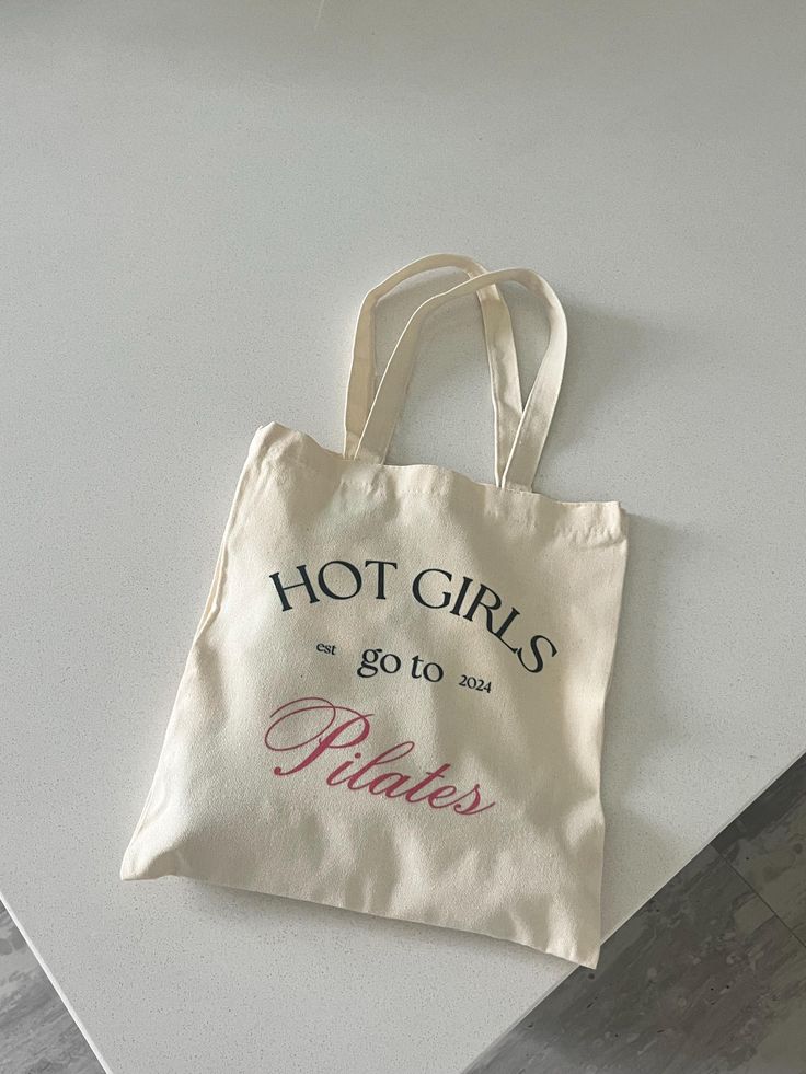 Canvas tote bag on a table with print: Hot Girls Go to Pilates 2024. Trendy fitness accessory for pilates enthusiasts.