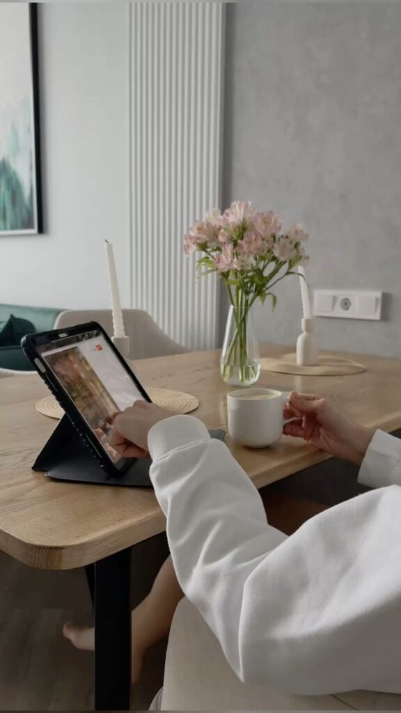 Person using a tablet at a wooden table with flowers and a mug, creating a cozy work-from-home atmosphere.