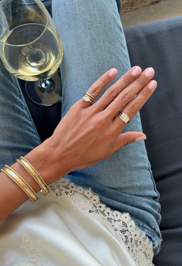 Elegant hand with gold rings and bracelets, holding wine glass. Casual jeans and lace top for a chic, sophisticated style.